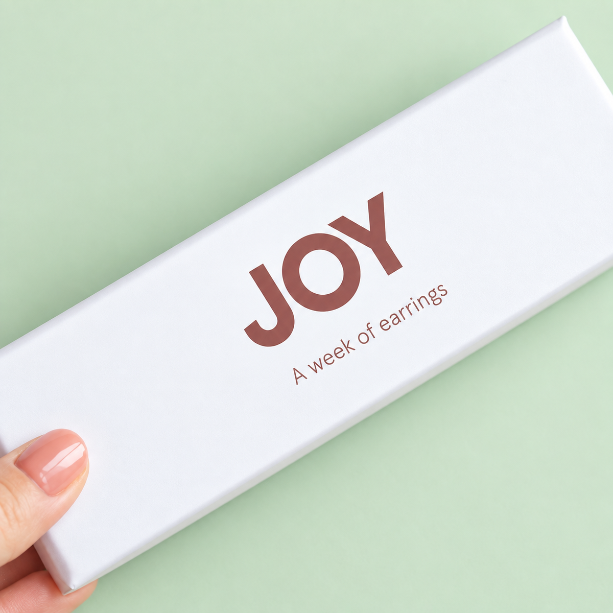 White JOY Week of Earrings gift box held against a soft pale green background.