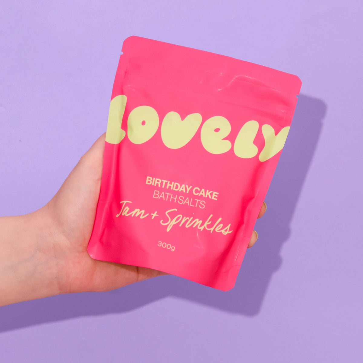 Hand holding a bright pink 300g pouch of Birthday Cake Bath Salts against a lilac background.
