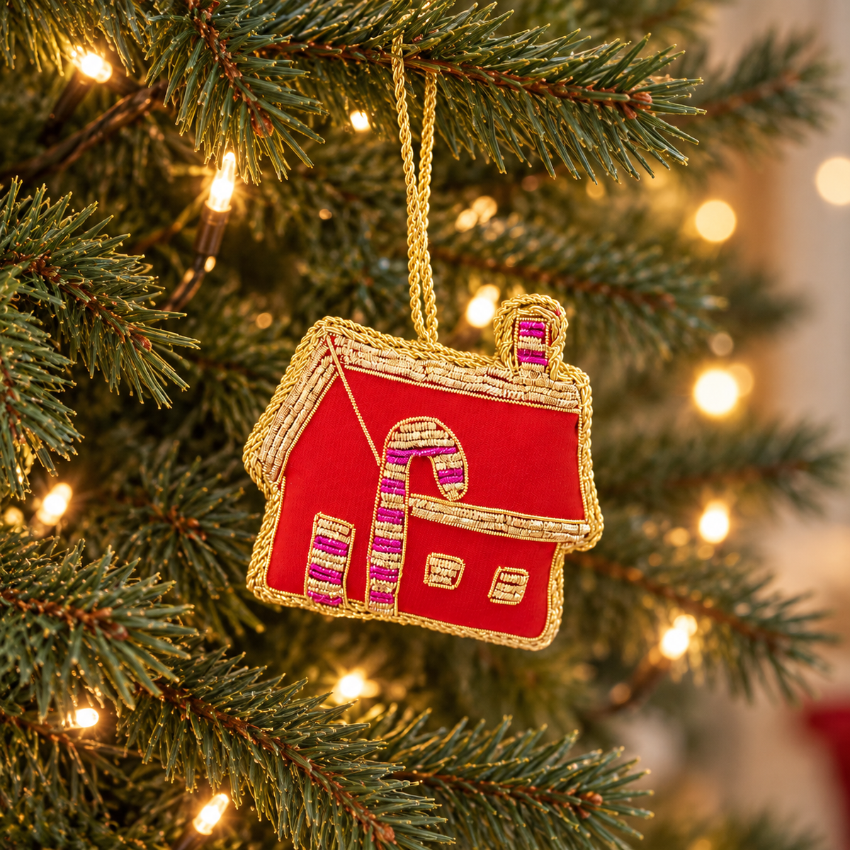 Red gingerbread house Christmas bauble with gold embroidery and pink candy cane details hanging on a lit Christmas tree.