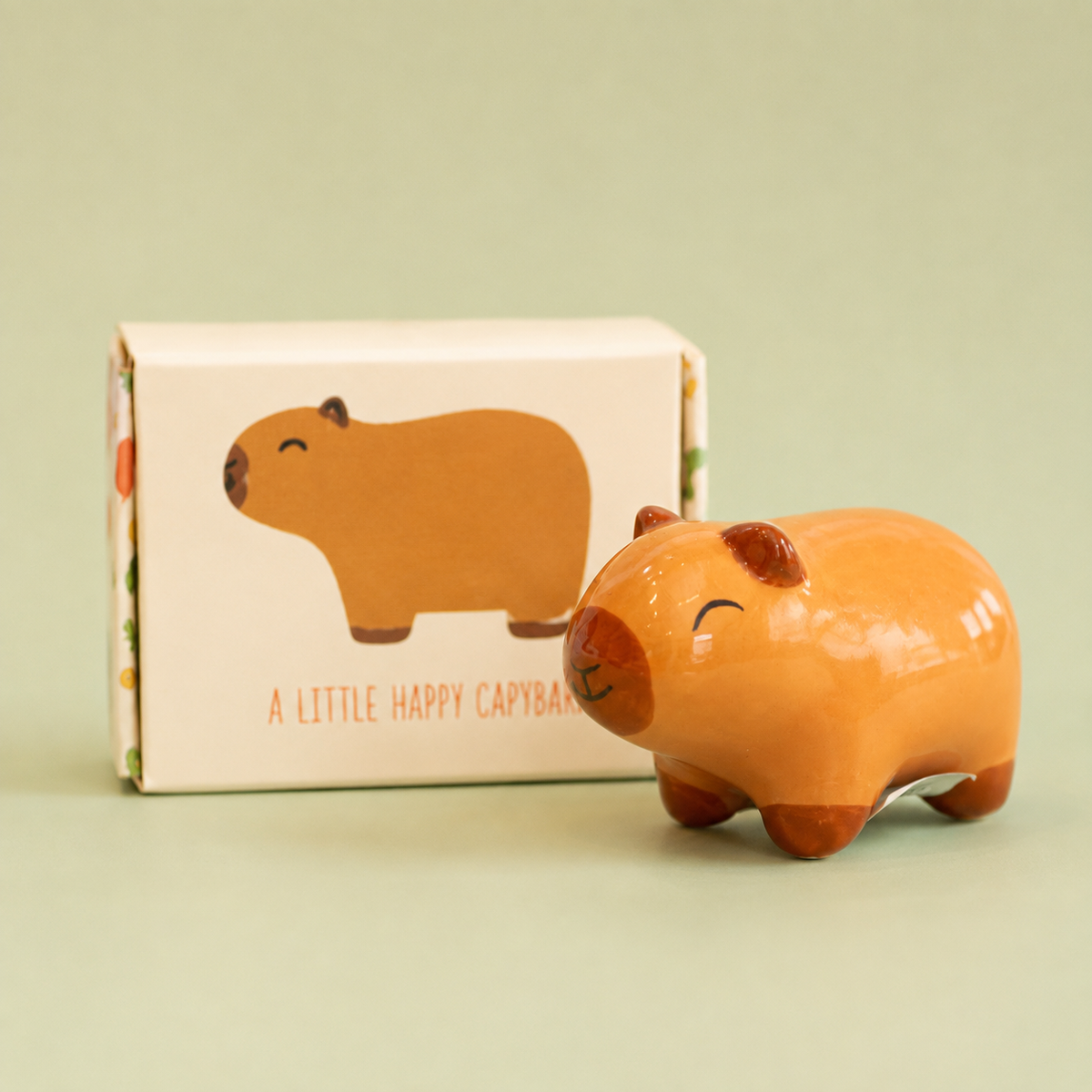 Little happy capybara ceramic token with smiling face beside its illustrated gift box on a soft green background.