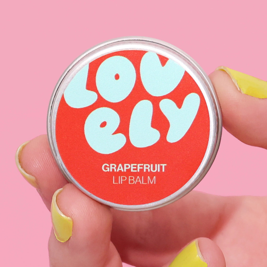 Grapefruit Lip Balm tin held in a hand against a pink background.
