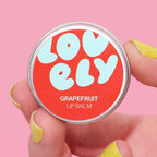 Grapefruit Lip Balm tin held in a hand against a pink background.
