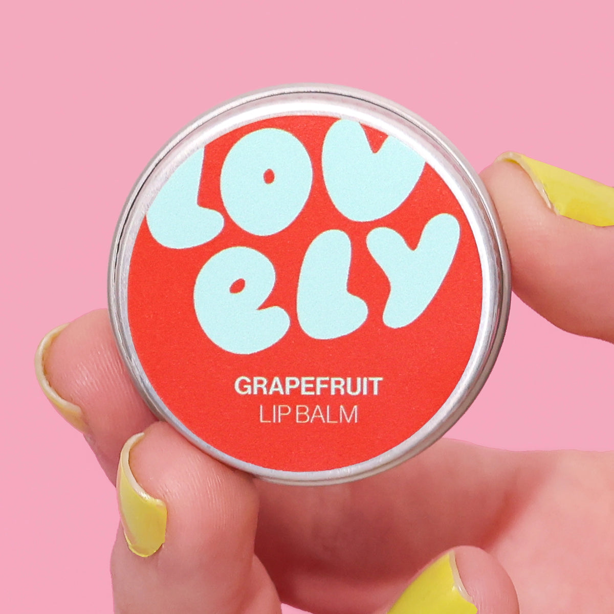 Grapefruit Lip Balm tin held in a hand against a pink background.

