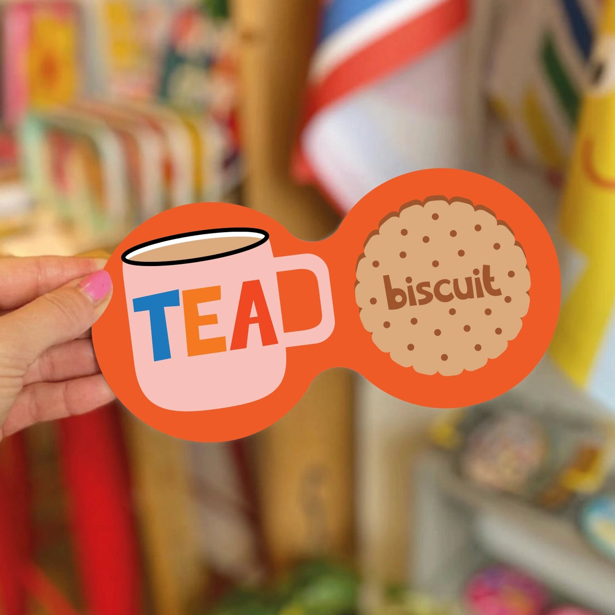 Bright orange double coaster with pink tea mug, colourful TEA lettering and illustrated biscuit, held inside a colourful shop.
