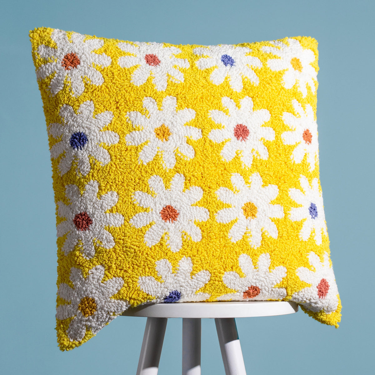 Yellow tufted cushion covered in white daisies with colourful centres, styled against a blue background.