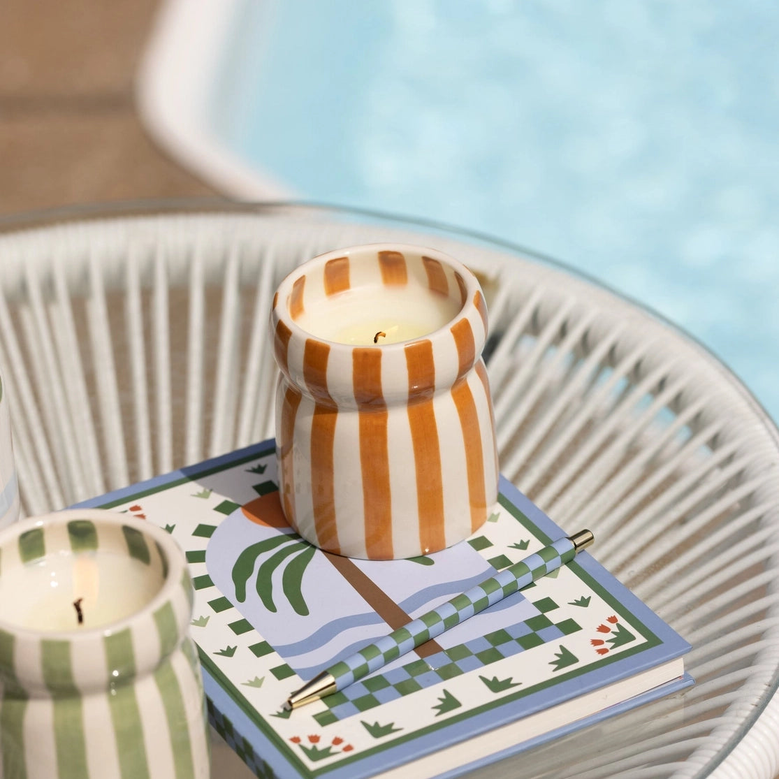 Lit Sienna Sunset striped ceramic candle styled on a colourful book beside a swimming pool.