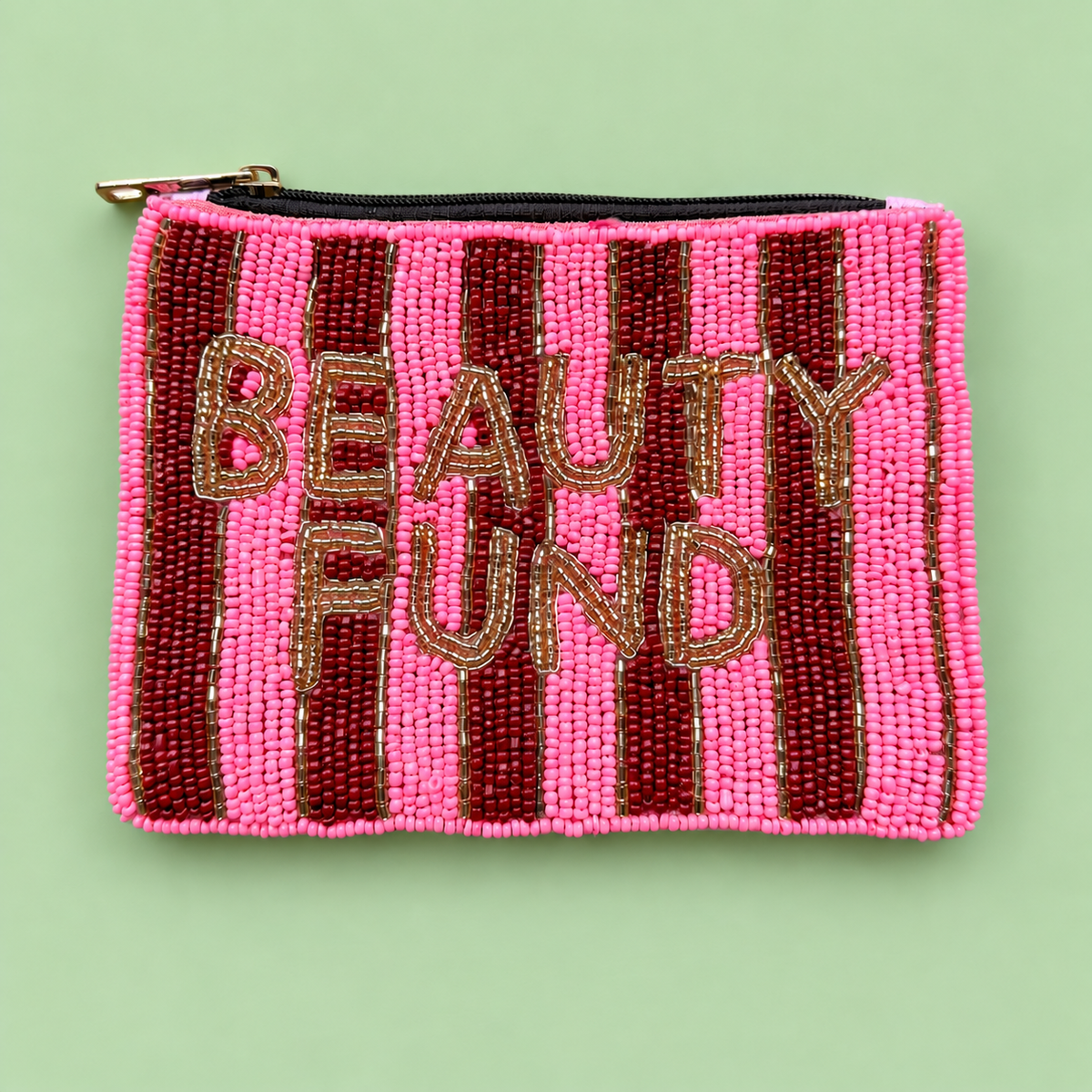 A rectangular beaded pouch with pink and dark red vertical stripes, featuring the words 'BEAUTY FUND' in gold beaded letters. The pouch has a black zipper with a gold pull tab and is photographed flat on a light green background.