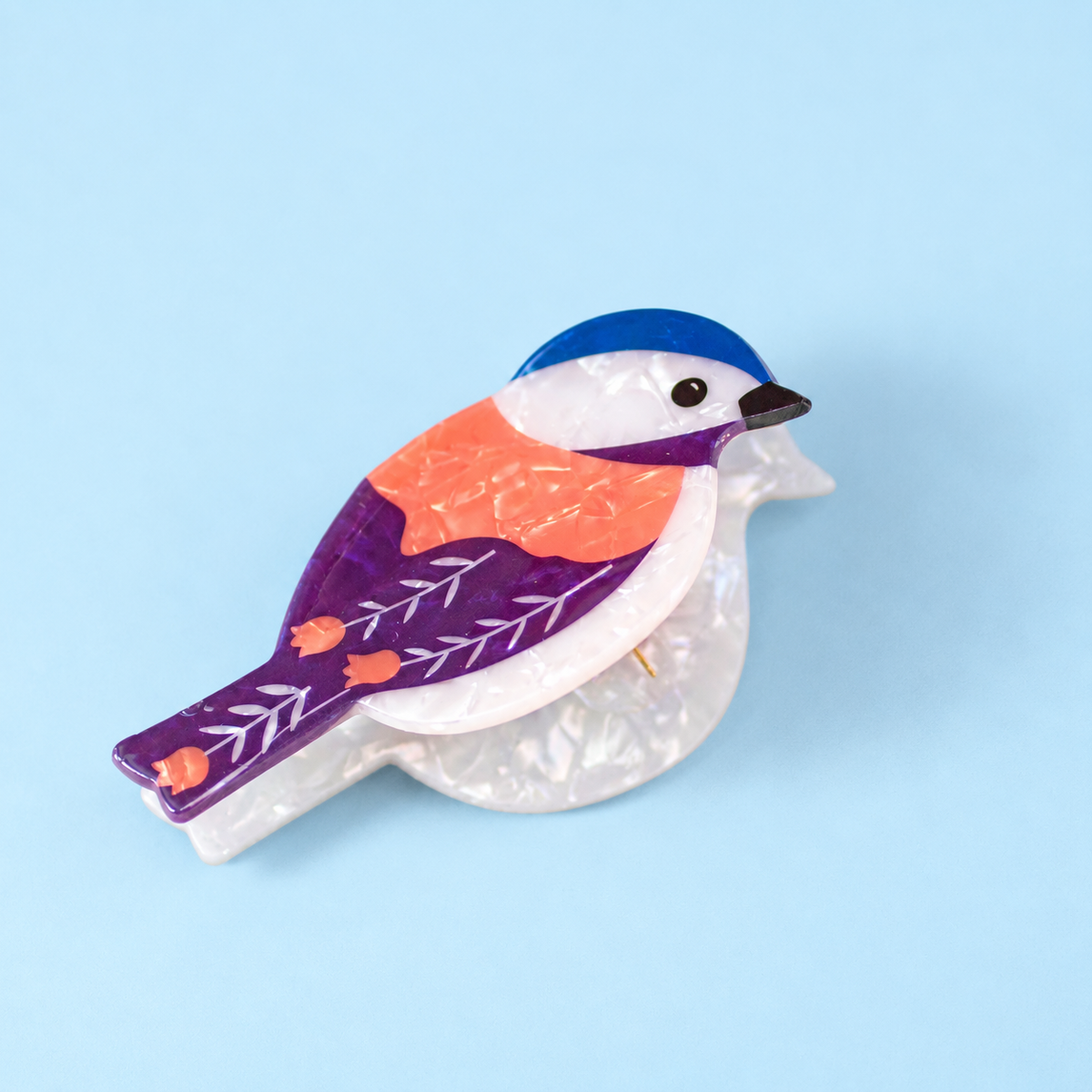 Colourful pearlescent bird hair claw clip in purple, coral, blue and white with floral wing details on a pale blue background.