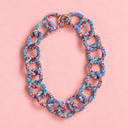 Chunky blue mix beaded statement chain necklace with multicoloured interlocking links and gold tone ring fastening on a soft pink background.