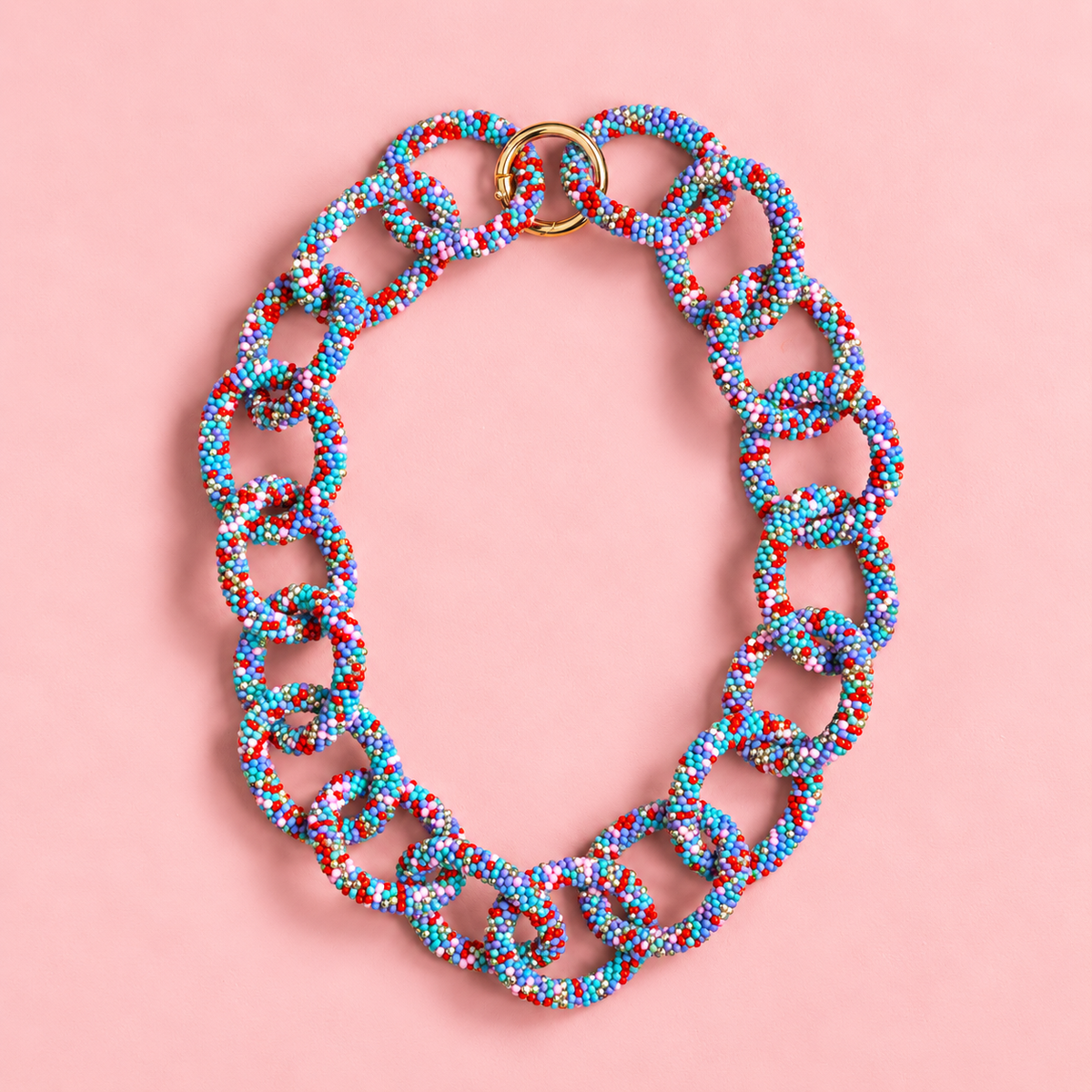 Chunky blue mix beaded statement chain necklace with multicoloured interlocking links and gold tone ring fastening on a soft pink background.