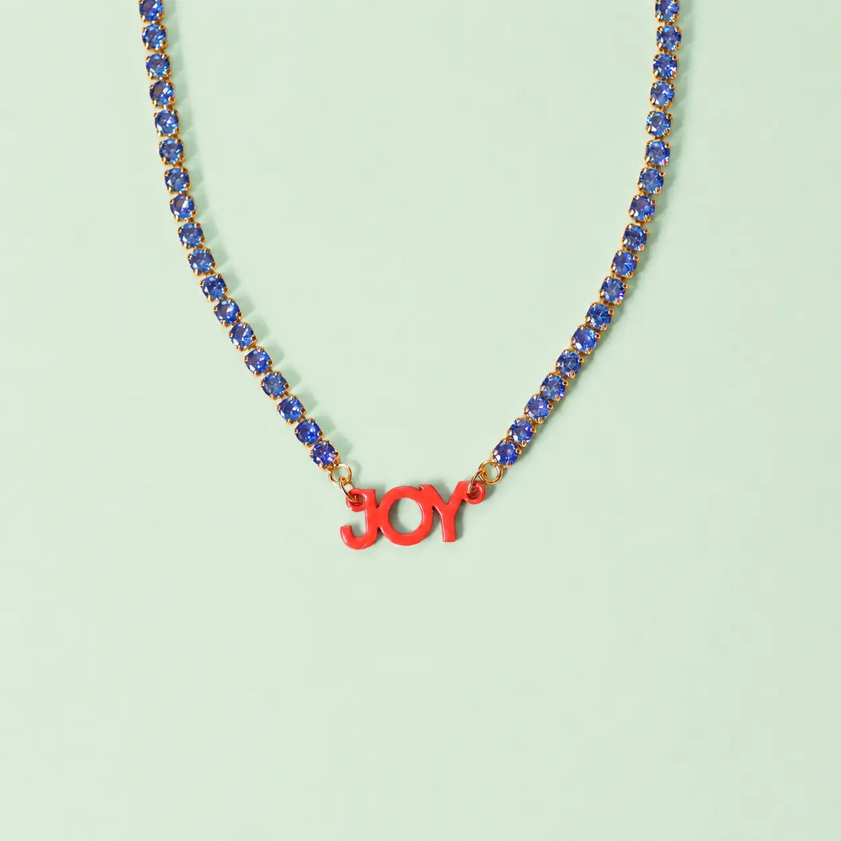 Pocketful of Joy signature necklace with a vibrant blue crystal tennis chain and coral JOY centrepiece on a mint green background.