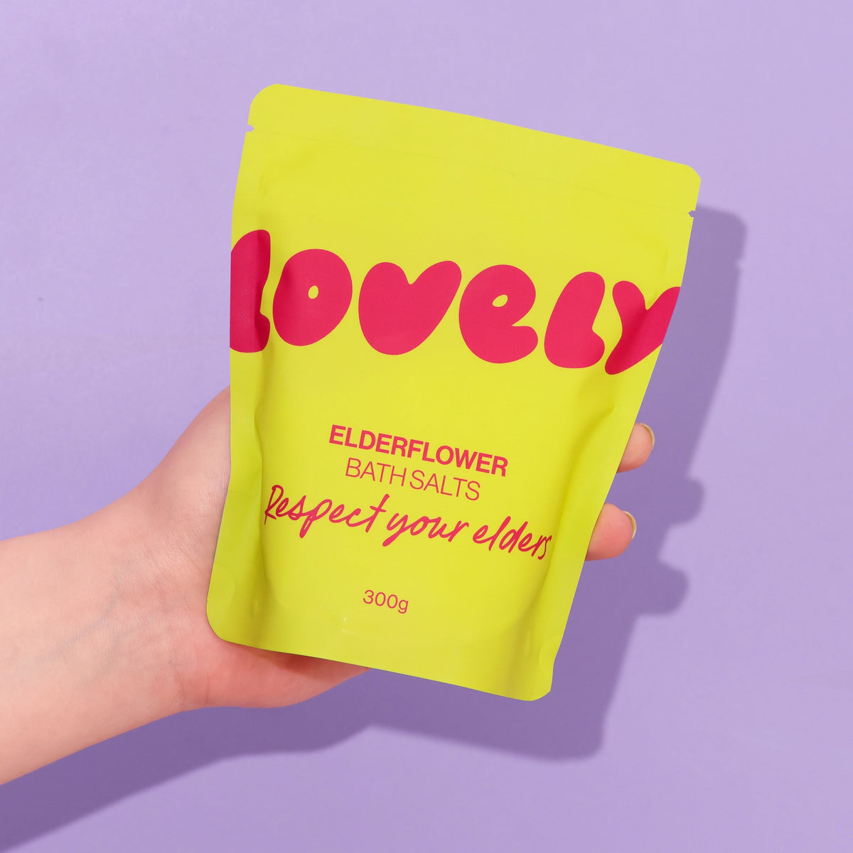 Elderflower Bath Salts in bright yellow Respect Your Elders packaging, held against a lilac background.