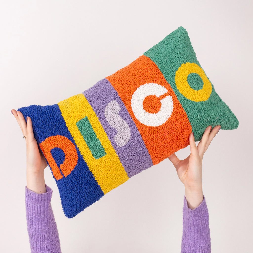 A colorful, chunky tufted pillow with the word 'DISCO' in large letters, held up by two hands wearing purple knit sleeves against a plain light background.
