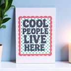 Cheerful Cool People Live Here A5 art print with bold retro lettering, green starbursts and a red scalloped border displayed on a shelf.