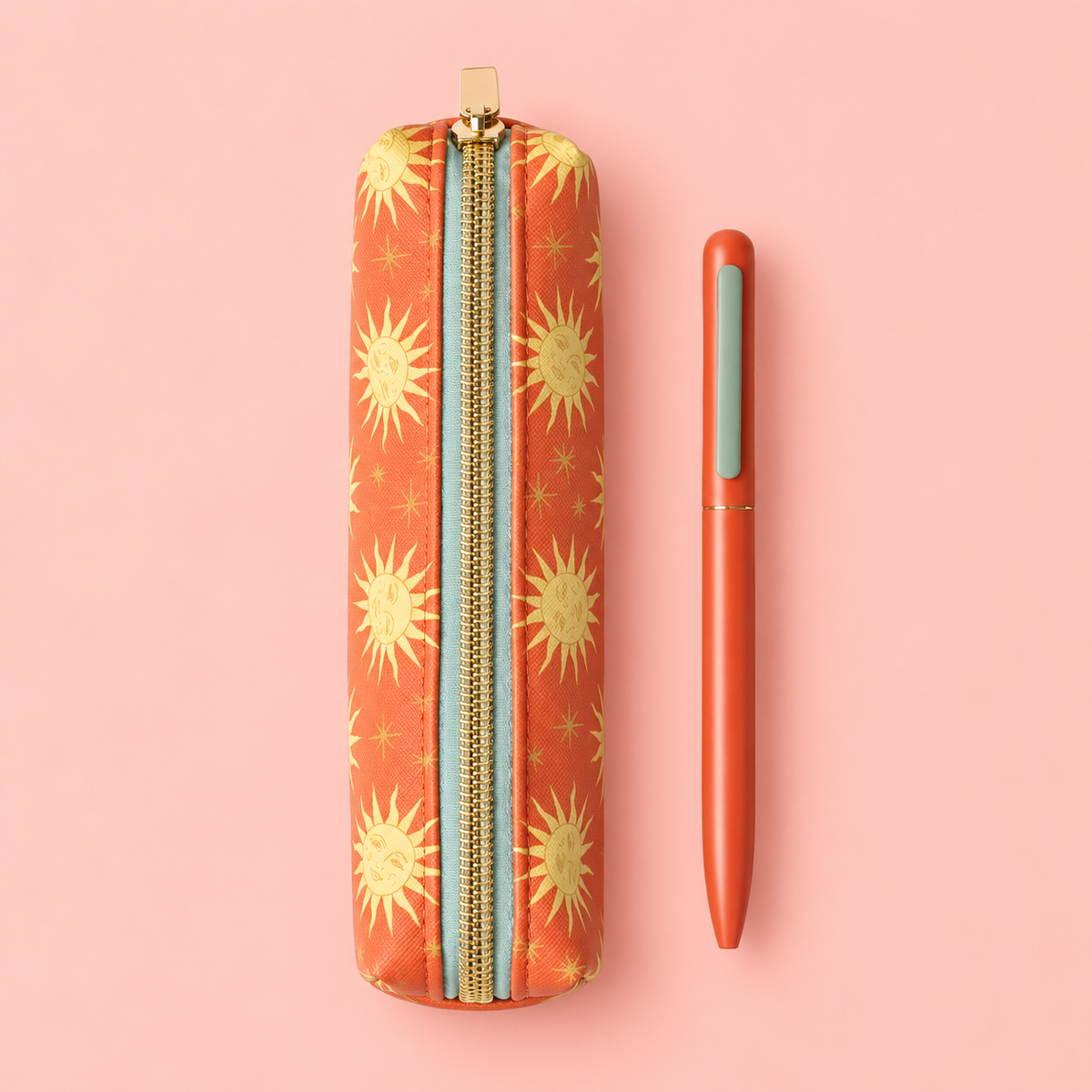 Terracotta and gold sun print pencil pouch with gold zip and coordinating orange pen on a pale pink background.