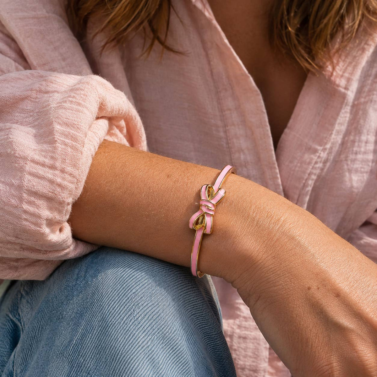 Pale pink enamel bow bangle with gold-tone detailing styled on the wrist with a pink blouse and denim.