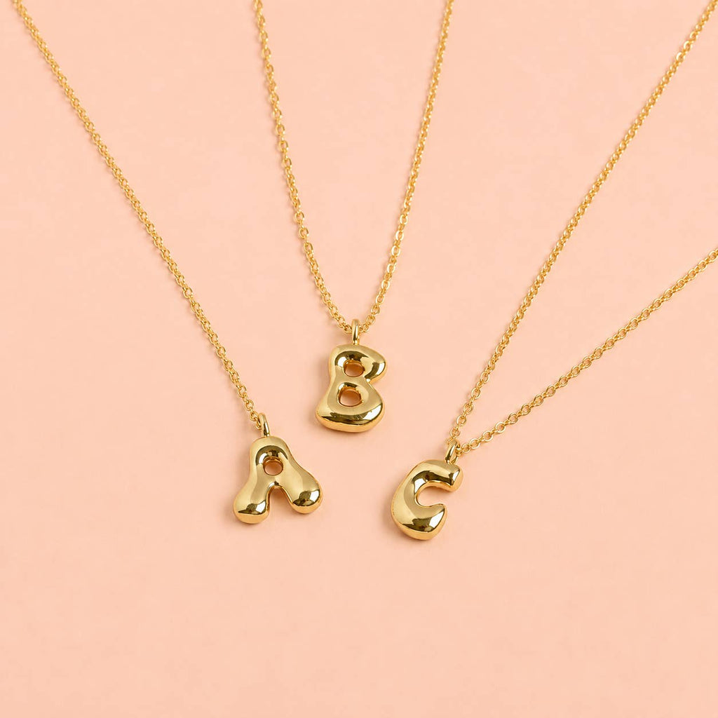 Gold bubble A, B and C initial necklaces with chunky rounded letter pendants displayed on a soft pink background.