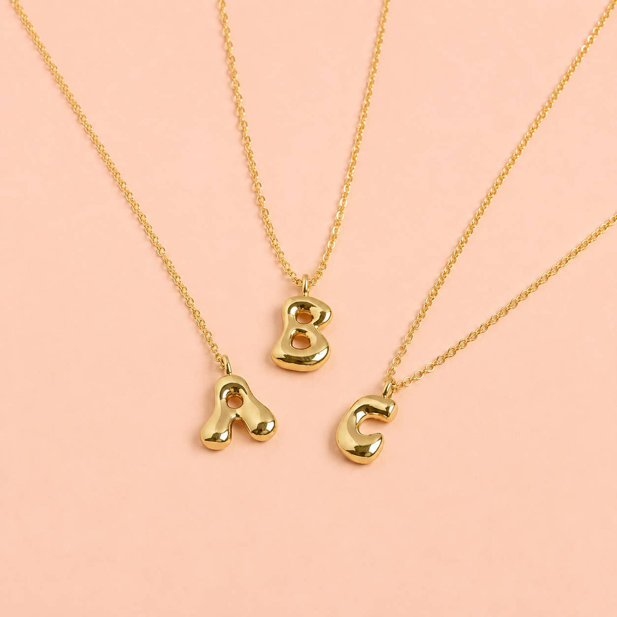 Gold bubble A, B and C initial necklaces with chunky rounded letter pendants displayed on a soft pink background.