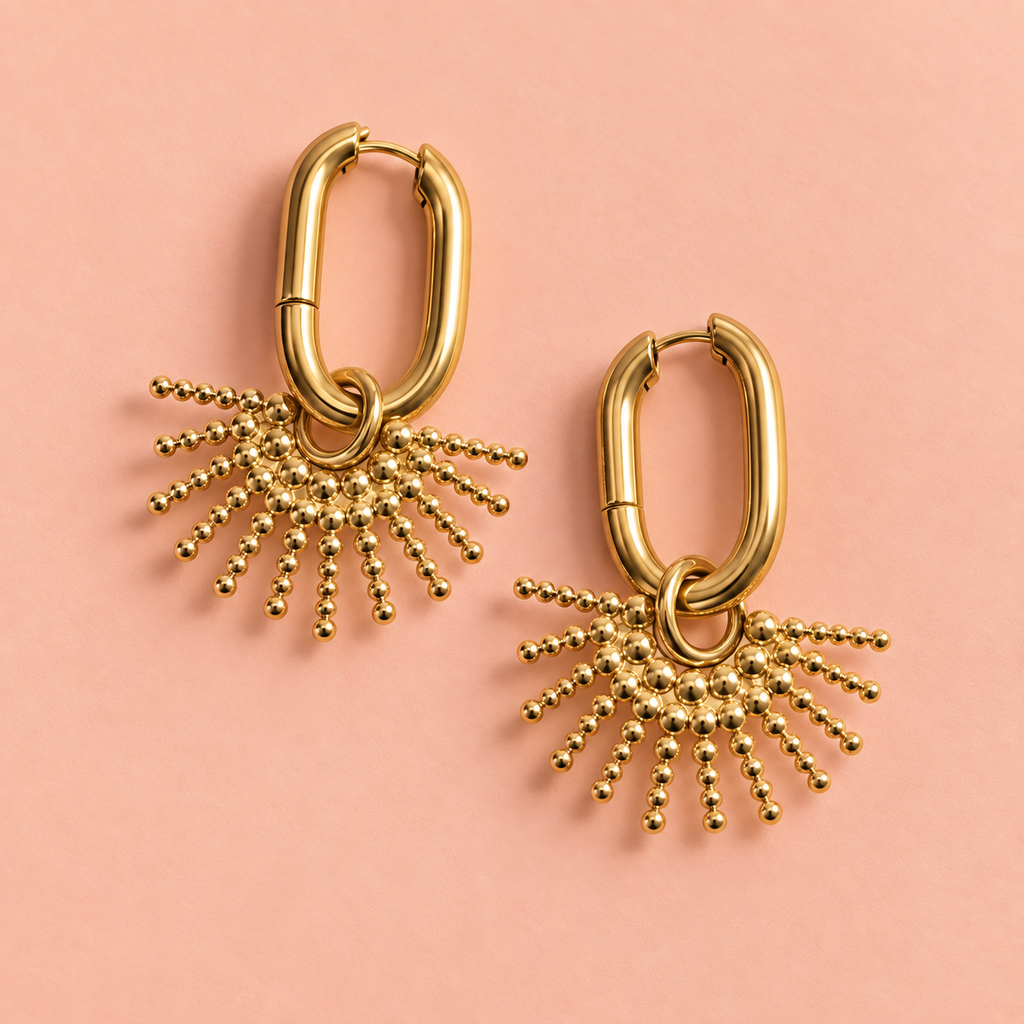 Gold sunburst drop hoop earrings lying on a soft peach background.