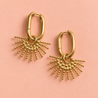 Gold sunburst drop hoop earrings lying on a soft peach background.