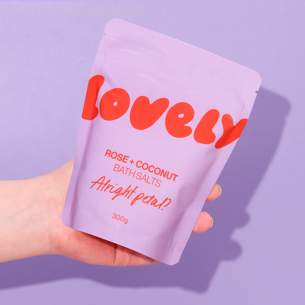 Rose and Coconut Bath Salts in lilac Alright Petal packaging, held against a purple background.