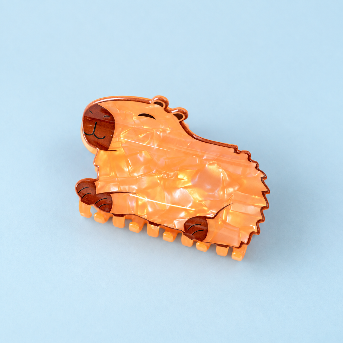 Pearlescent orange capybara shaped hair claw clip with cute face and brown details on a soft pale blue background.