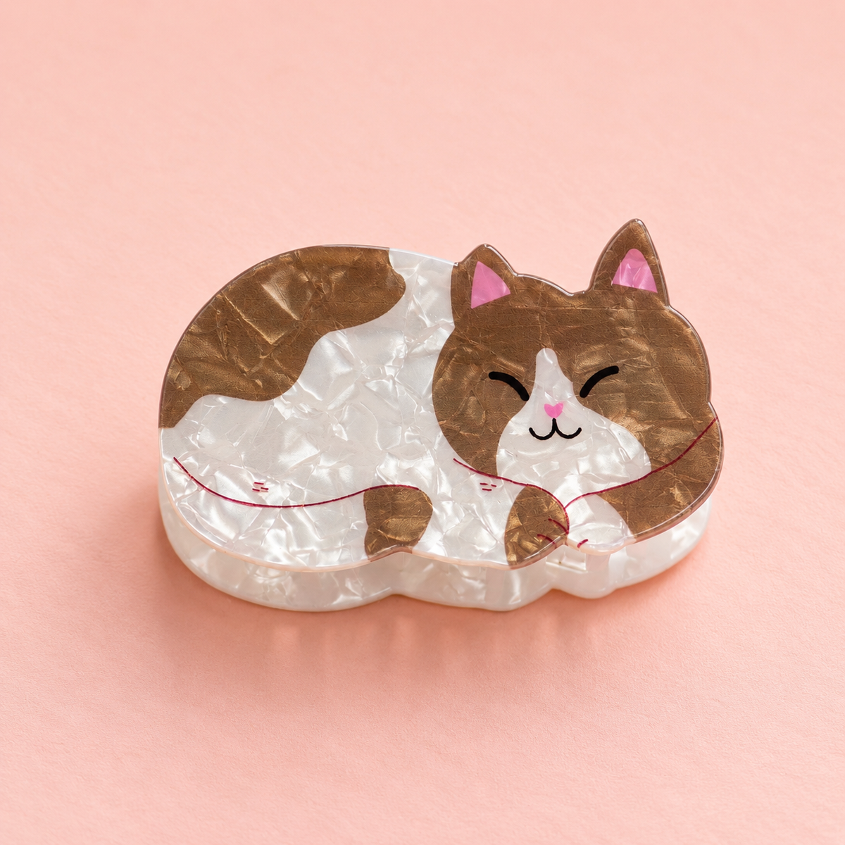 Curled up cat shaped acrylic claw hair clip in pearlescent white and warm brown with pink details on a soft peach background.