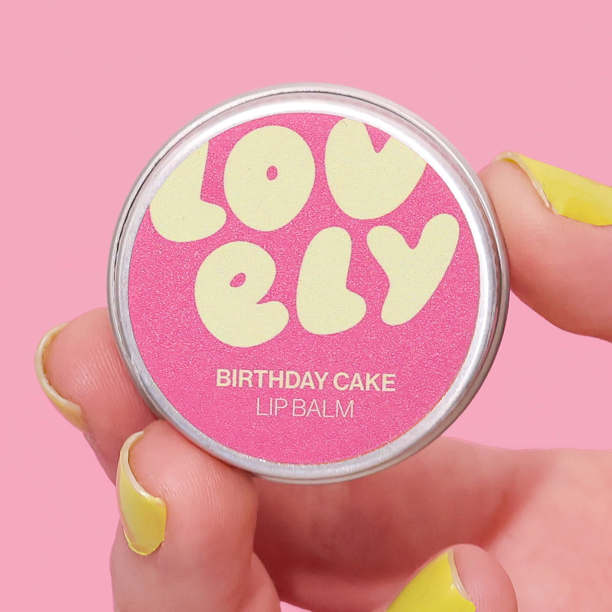 Birthday Cake Lip Balm tin held in a hand against a pink background.
