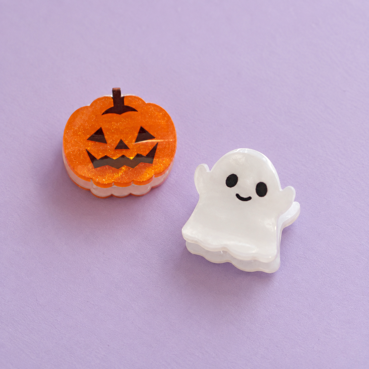 Mismatched mini Halloween hair clips featuring a sparkly orange pumpkin and pearly white ghost on a lilac background.
