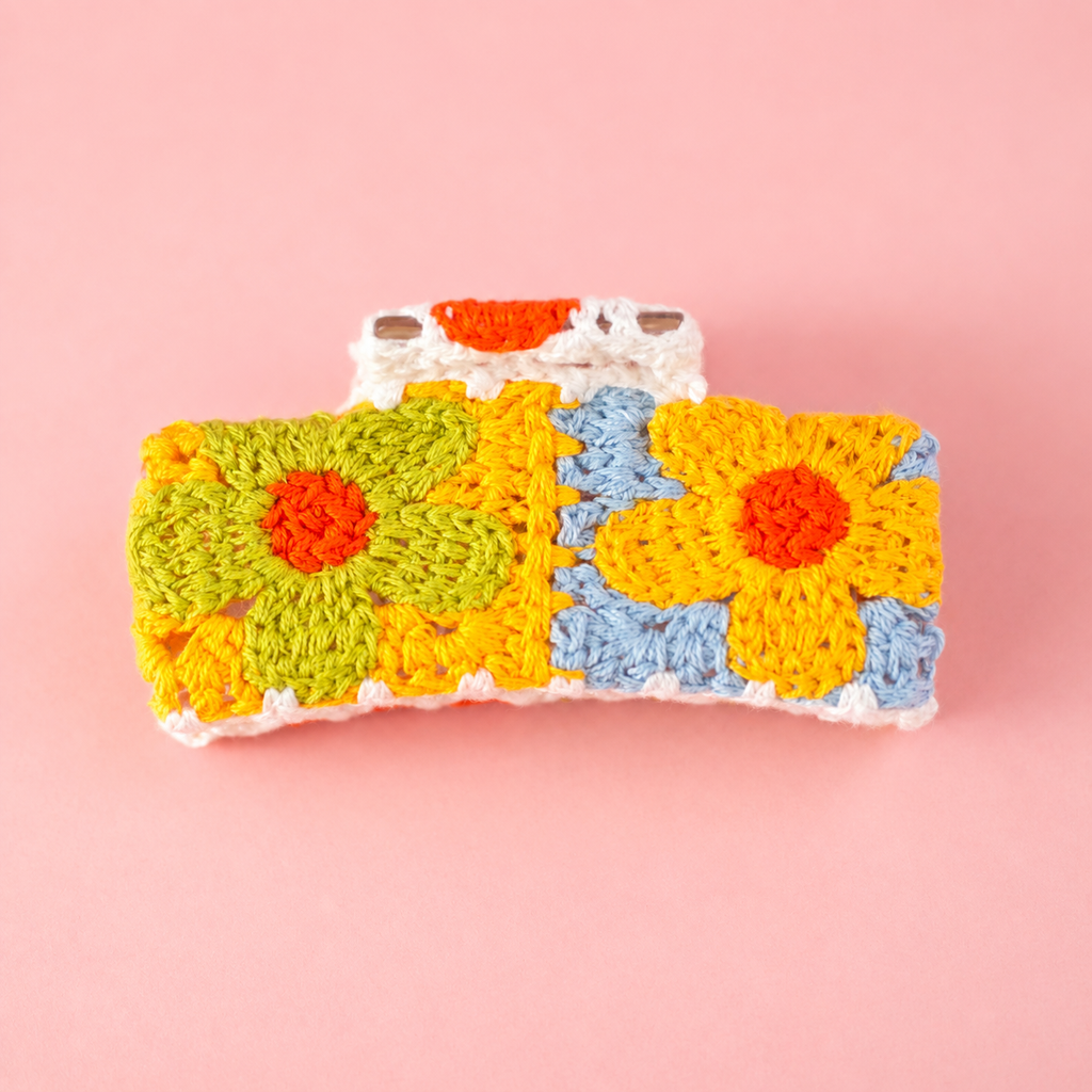 Colourful crochet flower claw hair clip in orange, yellow, green, blue and cream on a soft pink background.
