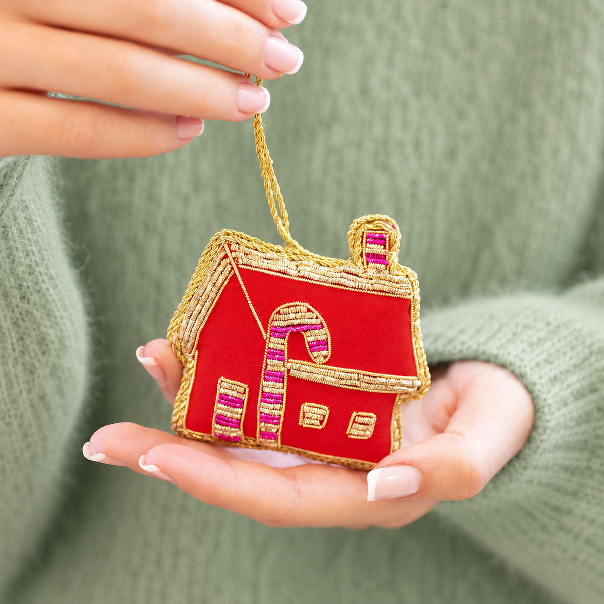 Red and gold embroidered gingerbread house Christmas bauble with pink details held in a hand.