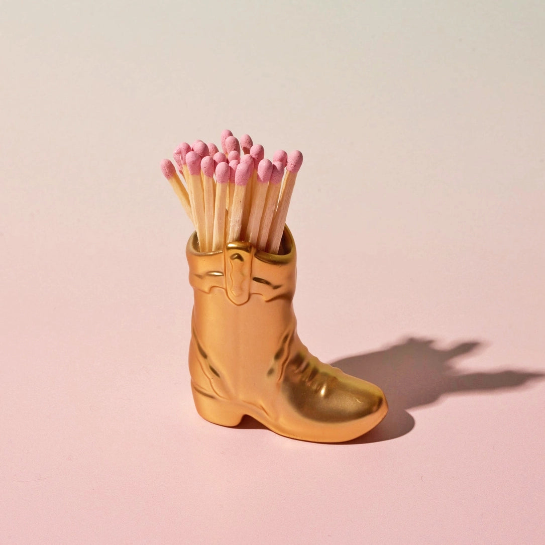 Gold cowboy boot match holder with pink tipped matches on a soft pale pink background.