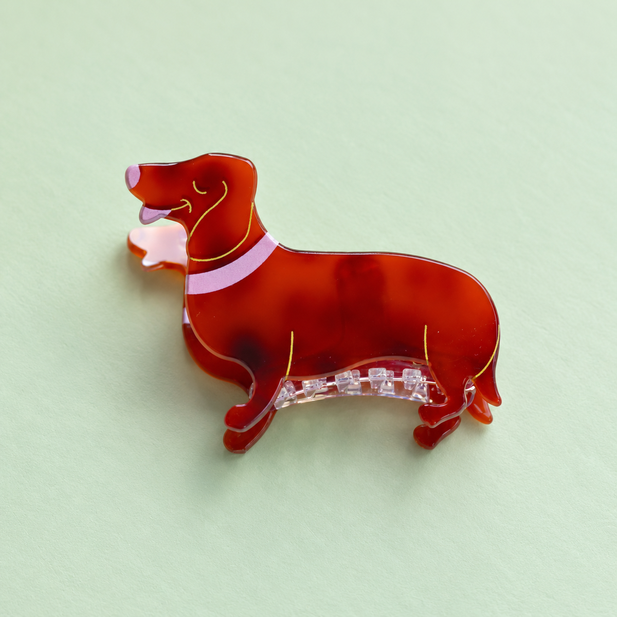 Reddish brown dachshund shaped acrylic claw hair clip with pink and gold details on a soft pale green background.