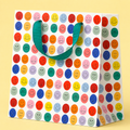 White gift bag covered in colourful smiling faces in pink, red, blue, green and yellow with a green ribbon handle.