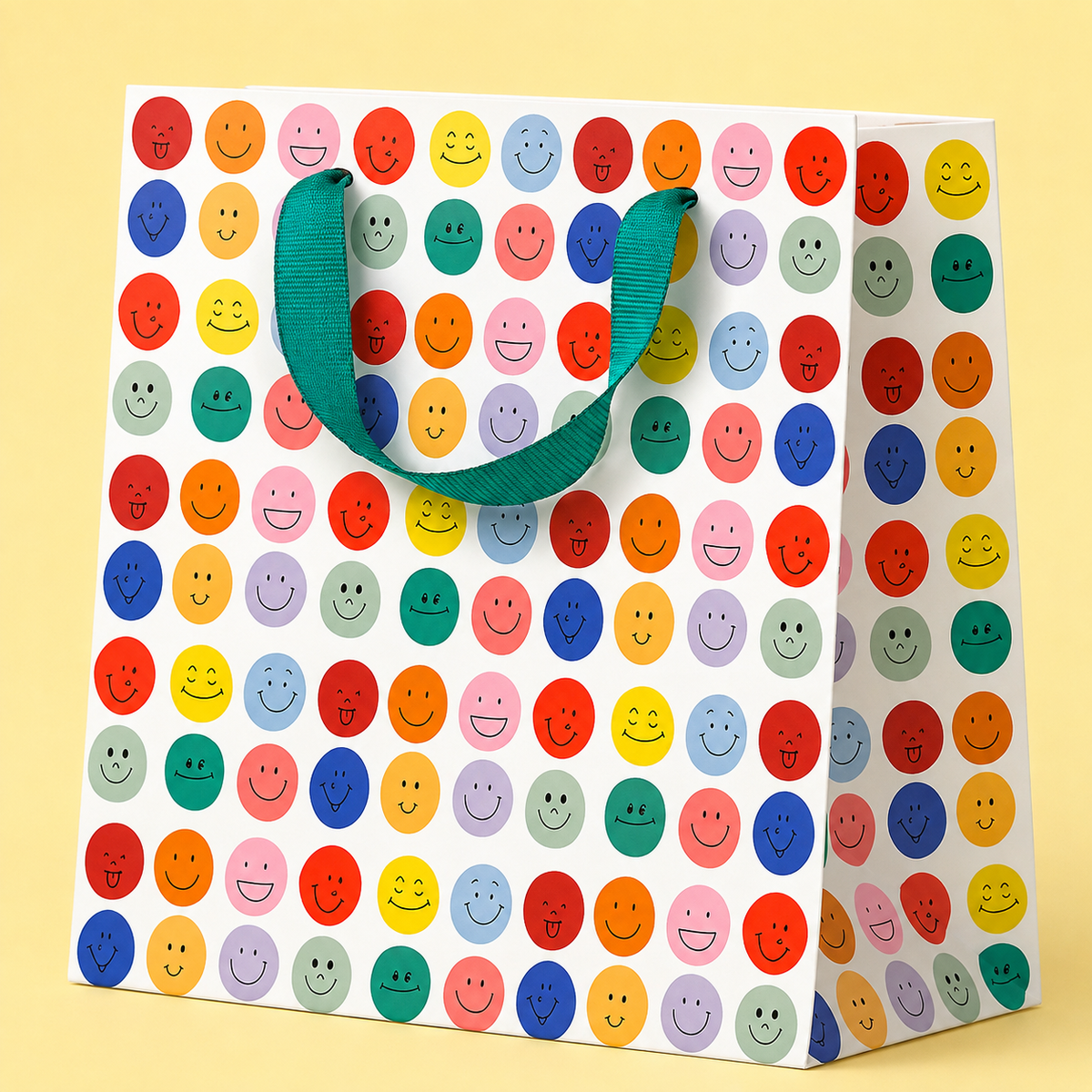 White gift bag covered in colourful smiling faces in pink, red, blue, green and yellow with a green ribbon handle.