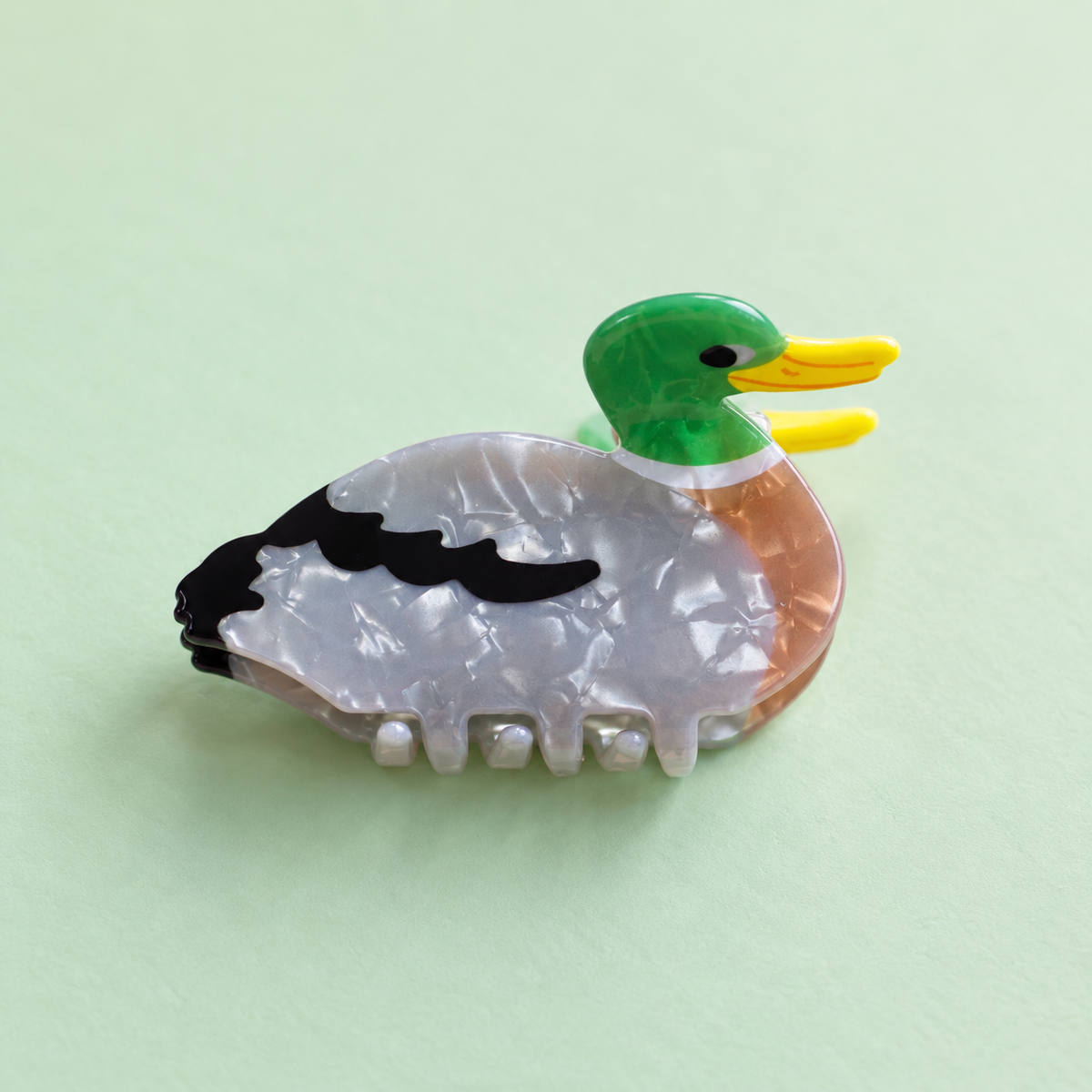 Colourful acrylic duck claw hair clip with green head, yellow beak and pearly grey body on a pale green background.