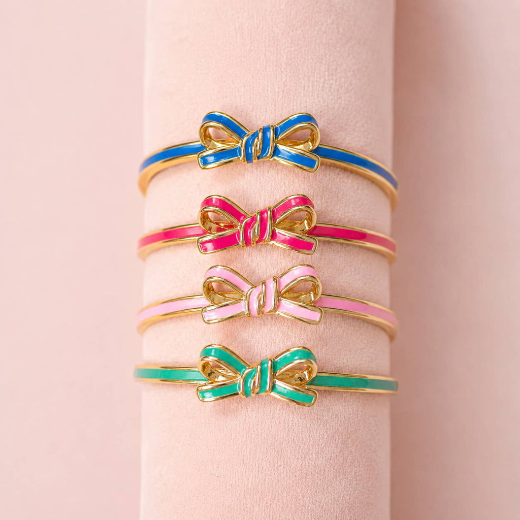 Four enamel bow bangles in blue, bright pink, pale pink and mint green displayed together on a blush jewellery stand.