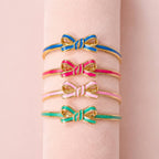 Four enamel bow bangles in blue, bright pink, pale pink and mint green displayed together on a blush jewellery stand.