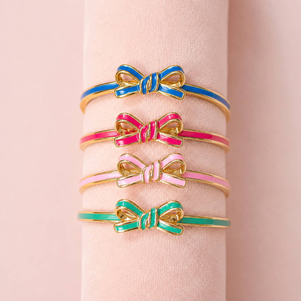 Four enamel bow bangles in blue, bright pink, pale pink and mint green displayed together on a blush jewellery stand.