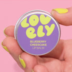 Blueberry Cheesecake Lip Balm tin held in a hand against a pink background.
