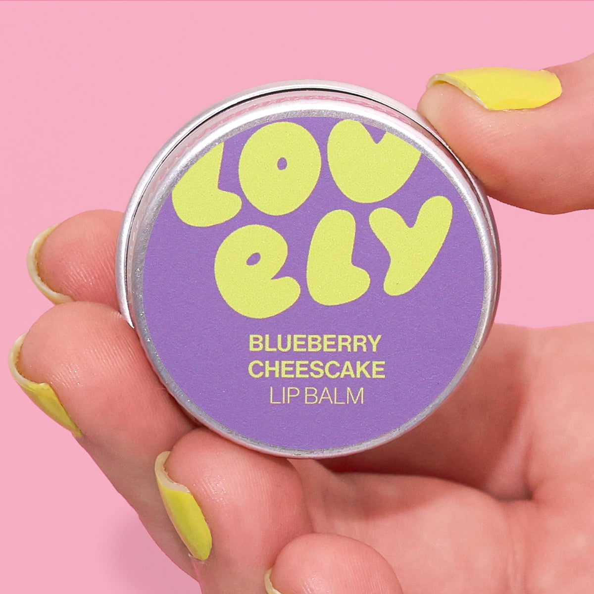 Blueberry Cheesecake Lip Balm tin held in a hand against a pink background.
