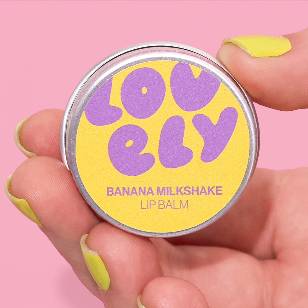 Banana Milkshake Lip Balm tin held in a hand against a pink background.
