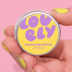 Banana Milkshake Lip Balm tin held in a hand against a pink background.
