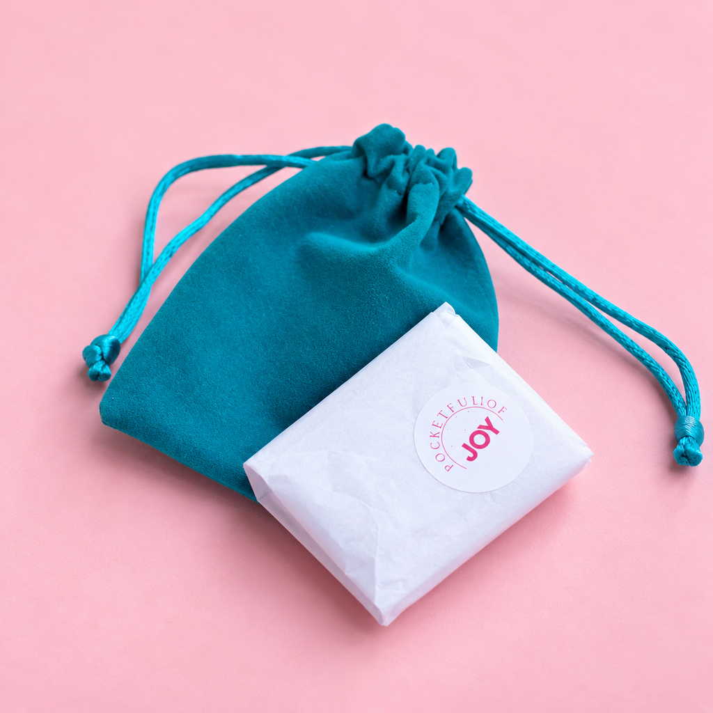A teal velvet drawstring pouch with a white paper-wrapped item featuring a circular 'Blossomjoy' logo, set against a solid pink background.