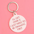 Pale pink round keyring with red text reading I’ll Get Over It, I Just Need To Be Dramatic First and a silver keyring.