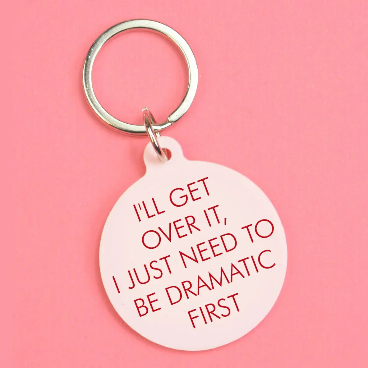 Pale pink round keyring with red text reading I’ll Get Over It, I Just Need To Be Dramatic First and a silver keyring.