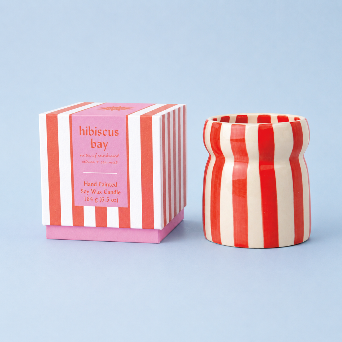 Hibiscus Bay soy wax candle beside its pink and red striped presentation box on a pale blue background.