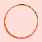 Orange dip enamel stacking bangle with polished gold tone detailing on a pale pink background.