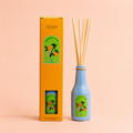 Orange Pop reed diffuser in a blue bottle with orange gift box and natural reeds on a soft peach background.