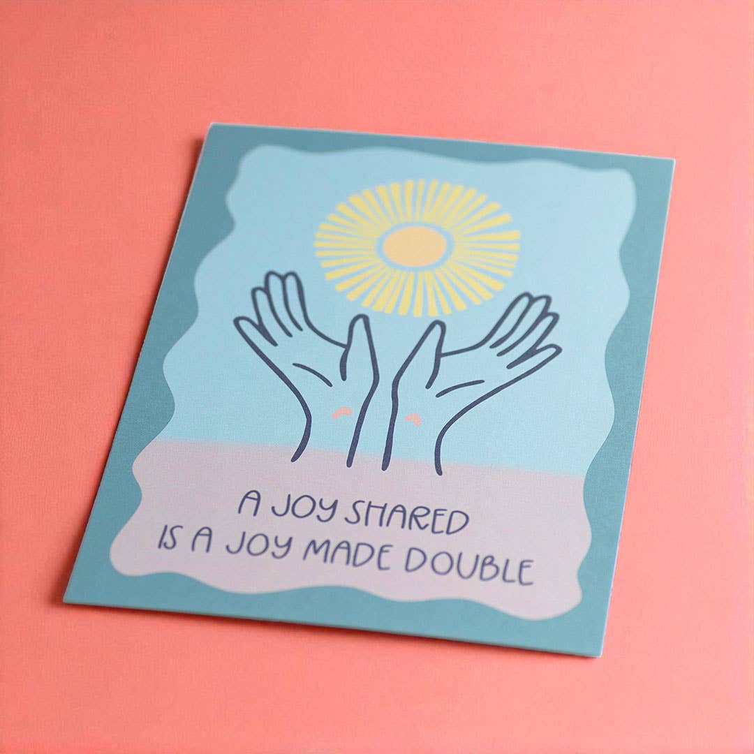 Uplifting A Joy Shared Is A Joy Made Double print in blue, pink and yellow, styled against a bright coral pink background.
